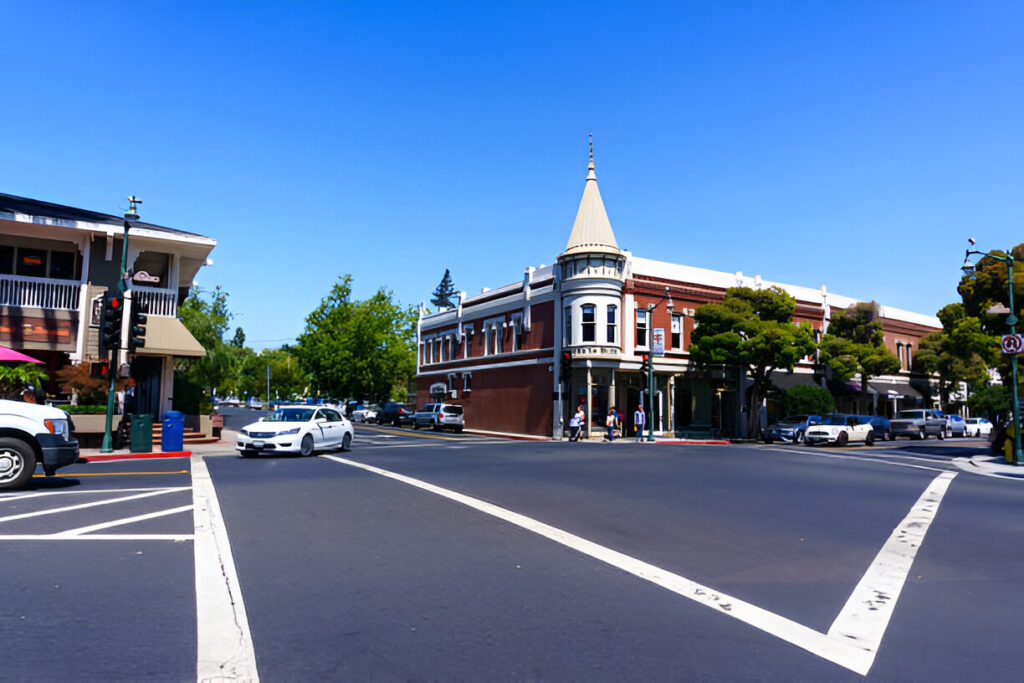 A lively street in Los Gatos, California, showing an intersection with cars, pedestrians crossing, trees, and traffic lights, capturing the town's charm and real estate appeal.